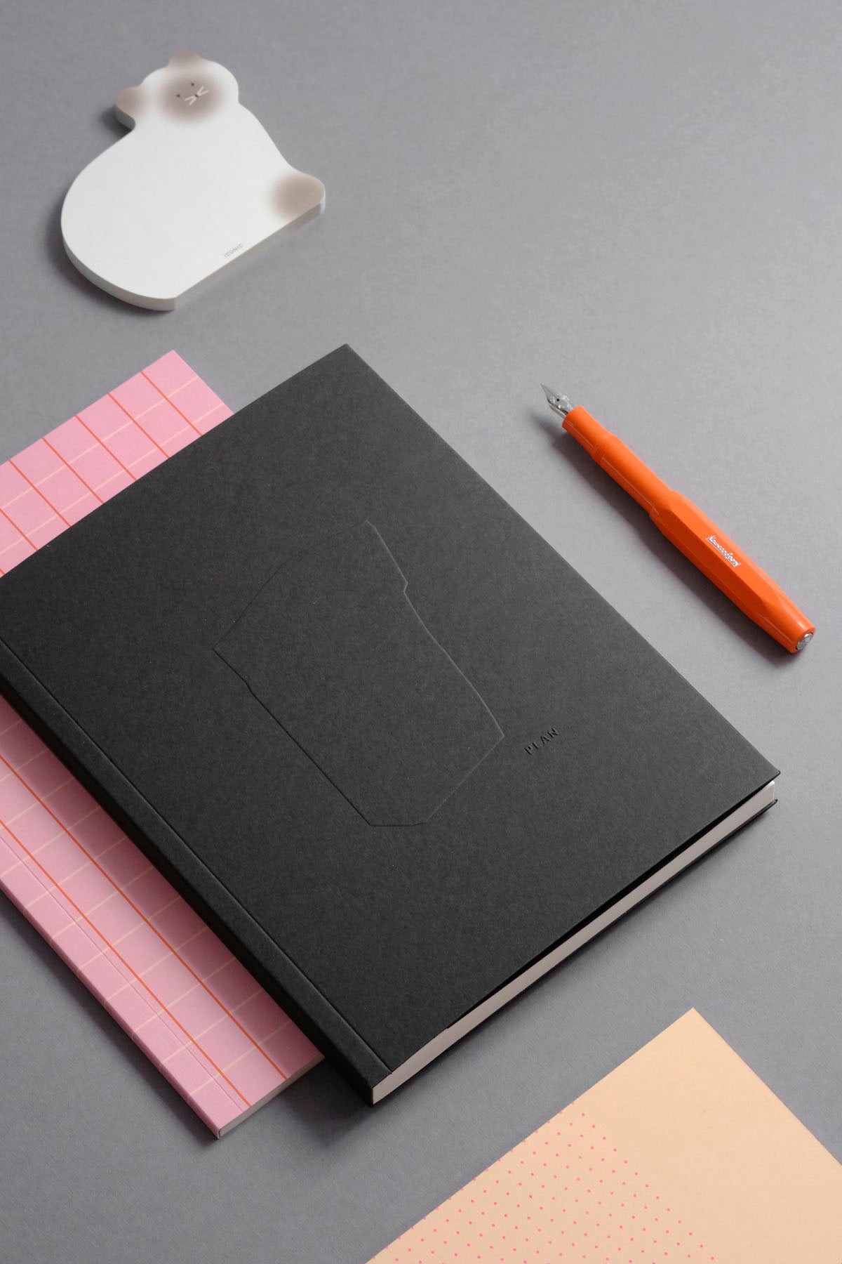 Planner Monolit w czarnej oprawie na tle różowego notesu Vita. Obok pióro wieczne Kaweco w kolorze pomarańczowym i wyrywane karteczki w kształcie kota.