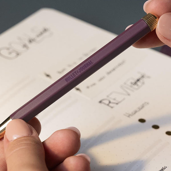 A plum-colored gel pen with the “Bullet Journal” inscription and gold details, held in hands.