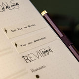 A dotted notebook with a plum-colored Drehgriffel gel pen attached with an elastic band.