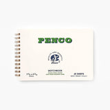 A cream-colored sketchbook with a metal spiral binding, featuring green lettering with the Penco brand name and smaller text containing information about the sketchbook.