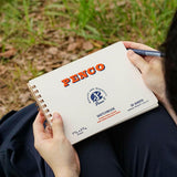 A cream-colored spiral-bound sketchbook with red lettering of the Penco brand name, held in hands.